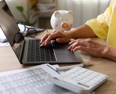 Frau arbeitet im Homeoffice am Laptop mit Taschenrechner und Gehaltsdaten auf dem Tisch