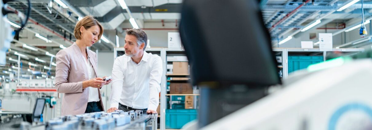 Zwei Fachkraefte in heller Kleidung im Gespraech neben einer Produktionslinie in einer modernen Industriehalle