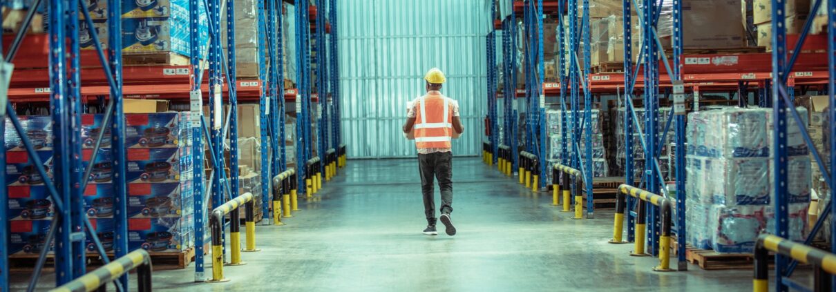 Arbeiter mit Warnweste läuft durch modernen Lagergang mit Regalsystemen | Hochregallager